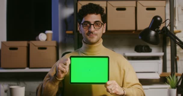 Um jovem olha diretamente para a câmera e mostra um tablet com tela verde — Vídeo de Stock