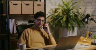 Genç adam cep telefonuyla konuşuyor ve ofiste dizüstü bilgisayar kullanıyor.