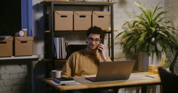 Joven hablando en un teléfono móvil y usando un portátil en la oficina — Vídeos de Stock