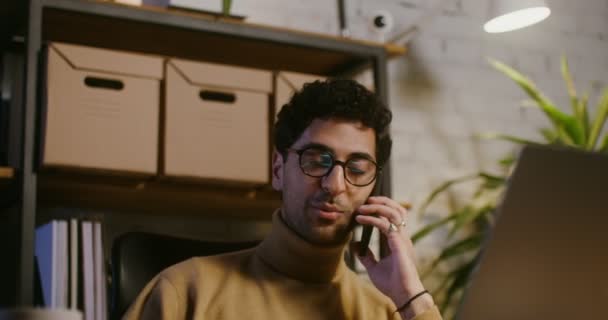Genç adam cep telefonuyla konuşuyor ve ofiste dizüstü bilgisayar kullanıyor. — Stok video