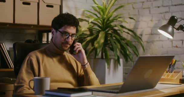 Joven hablando en un teléfono móvil y usando un portátil en la oficina — Vídeo de stock