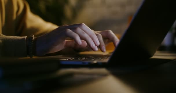 Joven hombre de piel clara está escribiendo en un ordenador portátil mientras está sentado junto a la luz de las lámparas — Vídeos de Stock