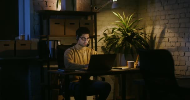Een jonge man typt op een laptop en praat in een microfoon in het kantoor — Stockvideo