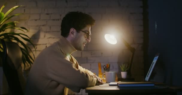 Un joven está hablando en un enlace de vídeo, sentado en una mesa frente a un ordenador portátil — Vídeos de Stock