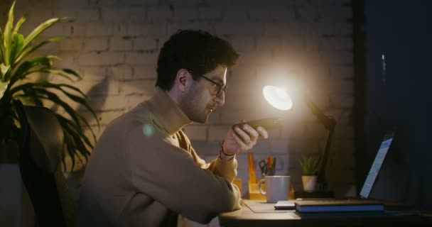 Fiatal férfi beszél kéz nélkül egy mobiltelefon, miközben ül a laptop előtt — Stock videók