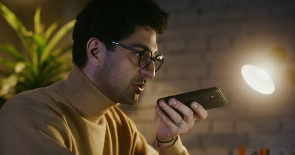 Joven hablando con las manos libres en un teléfono móvil mientras está sentado frente a la computadora portátil — Vídeos de Stock