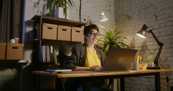 Mujer joven sonriendo mirando directamente a la cámara sentada frente a la computadora portátil — Vídeos de Stock