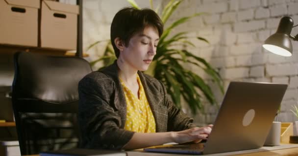 Een jonge vrouw met een serieuze blik typt op een laptop in een modern kantoor — Stockvideo