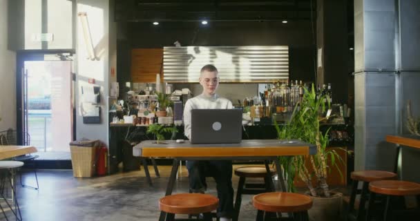 Young trabaja remotamente en una cafetería escribiendo en un portátil y bebiendo café — Vídeos de Stock