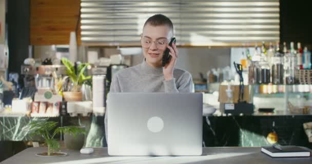 Egy fiatal nő felveszi a telefont, felnéz a munkából egy laptopon. — Stock videók