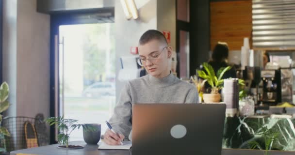 Mujer joven hace notas en el cuaderno sentado frente a la computadora portátil en la cafetería — Vídeo de stock