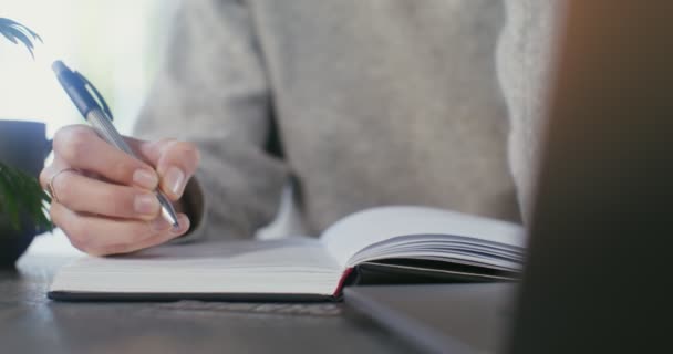 Mujer joven hace notas en el cuaderno sentado frente a la computadora portátil en la cafetería — Vídeo de stock