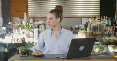 Genç bir kadın bilgisayarla konuşuyor, kafede bir masada oturuyor.