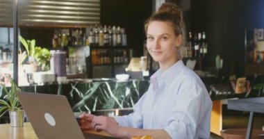 Kameraya bakıp gülümseyen bir kadın kafede dizüstü bilgisayarın önünde oturuyor.