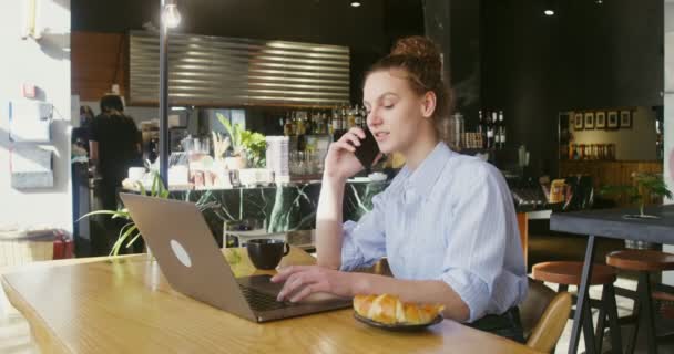 Junge Frau im Gespräch mit einem Handy, das vor einem Laptop in einem Café sitzt — Stockvideo