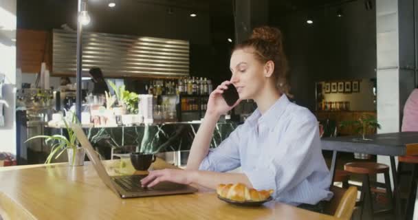 Mujer joven hablando en un móvil sentado frente a la computadora portátil en una cafetería — Vídeos de Stock