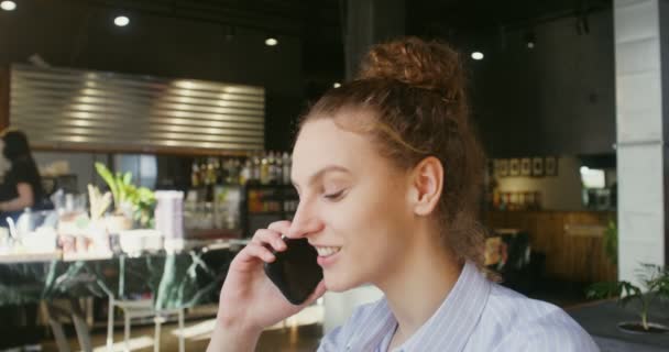 Eine junge Frau lächelt, während sie mit einem Mobiltelefon telefoniert, und sitzt im modernen Kaffee — Stockvideo