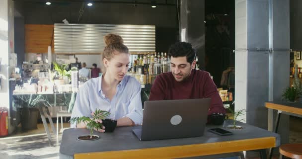Νεαρό ζευγάρι χαμογελά και να μιλάμε, ενώ χρησιμοποιώντας ένα φορητό υπολογιστή σε ένα σύγχρονο άνετο καφέ — Αρχείο Βίντεο