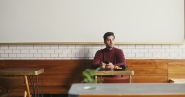 Genç bir adam bir kafede oturur ve zaman zaman telefon kullanır.