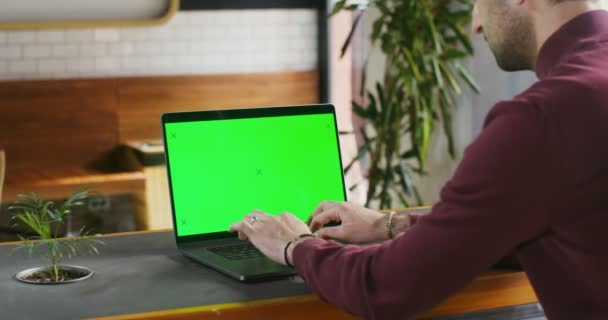 Un joven está escribiendo en un portátil con una pantalla verde sentado en una cafetería — Vídeos de Stock