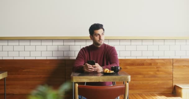 Молодой человек сидит в кафе и пользуется телефоном, время от времени оглядываясь вокруг — стоковое видео