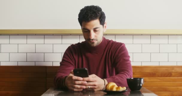 Een jongeman zit in een café en gebruikt af en toe telefoon om rond te kijken. — Stockvideo