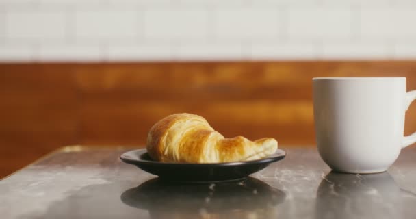 Ένα ορεκτικό κρουασάν σε ένα πιάτο και μια κούπα καφέ είναι σε ένα μικρό τραπέζι — Αρχείο Βίντεο