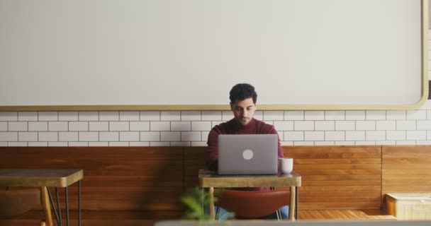 Junger Mann sitzt mit Laptop an kleinem Tisch in Café und arbeitet aus der Ferne — Stockvideo