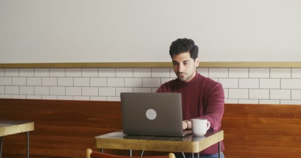 Jongeman gebruikt een laptop zittend aan een kleine tafel in een café, op afstand werkend — Stockvideo