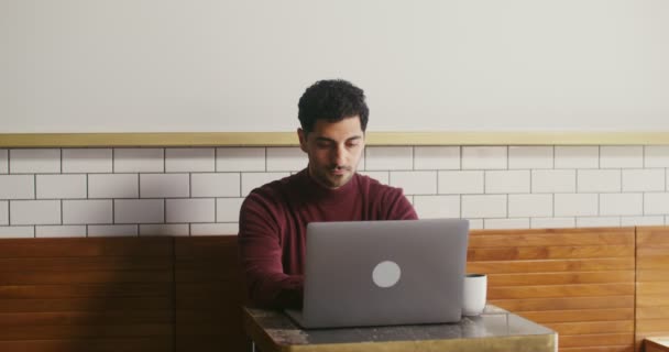 Jongeman gebruikt een laptop zittend aan een kleine tafel in een café, op afstand werkend — Stockvideo