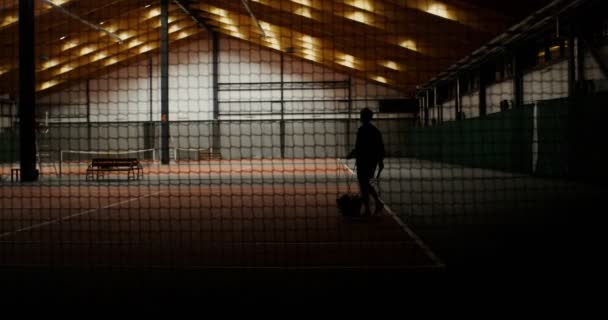 Un hombre entra en la cancha de tenis, llevando una canasta llena de pelotas de tenis — Vídeo de stock