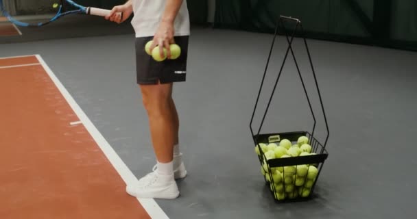 Een man pakt tennisballen uit een mand en slaat ze afwisselend met een racket — Stockvideo