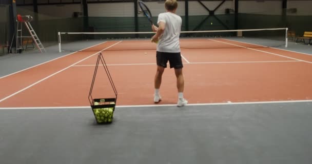 Un hombre entrena en una cancha de tenis solo, lanzando pelotas de tenis sobre la red — Vídeos de Stock