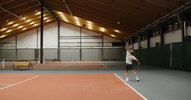 Un uomo inizia un set da tennis, colpendo la palla sul pavimento prima di lanciare — Video Stock