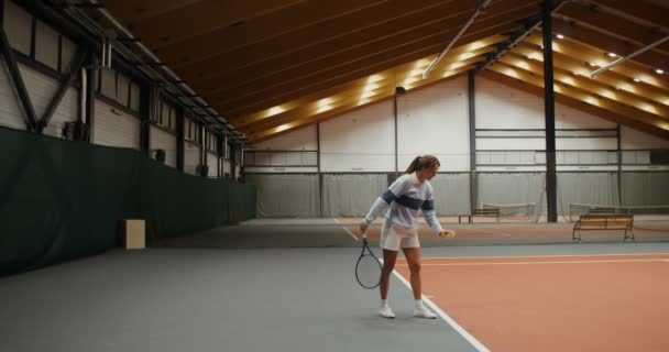 Una mujer comienza un set de tenis golpeando la pelota jugando en pista de tenis cubierta — Vídeos de Stock