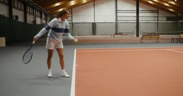 Una mujer comienza un set de tenis golpeando la pelota jugando en pista de tenis cubierta — Vídeos de Stock