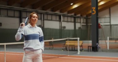 Genç bayan tenisçi tenis kortunda kameraya bakıp gülümsüyor.