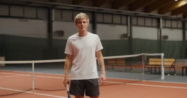 Genç adam gülümsüyor, tenis raketiyle poz veriyor, tenis kortunda dikiliyor.