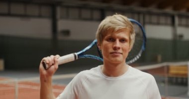 Genç adam gülümsüyor, tenis raketiyle poz veriyor, tenis kortunda dikiliyor.