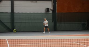 Genç bir adam uzun bir tenis takımına başlıyor, aktif olarak hareket ediyor ve topa vuruyor.