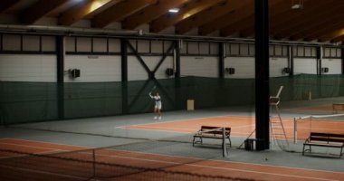 Bir kadın, büyük ve boş bir spor salonunun kortlarından birinde partneriyle tenis oynuyor.