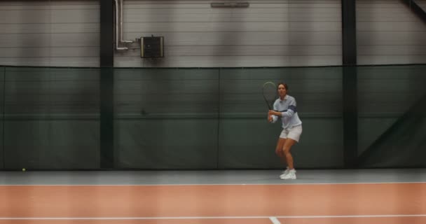 Una joven jugadora de tenis corre en la cancha de tenis golpeando una pelota de tenis — Vídeos de Stock