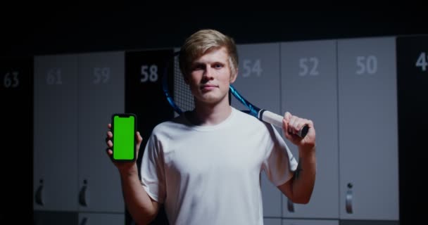 Een man met racket in zijn hand houdt telefoon vast met een groen scherm in de kleedkamer — Stockvideo