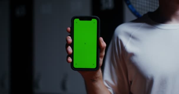 Een man met racket in zijn hand houdt telefoon vast met een groen scherm in de kleedkamer — Stockvideo