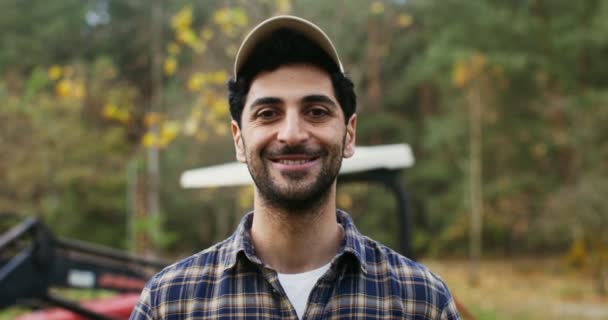 Un hombre sonriendo mirando a la cámara mientras está de pie contra la máquina agrícola — Vídeo de stock