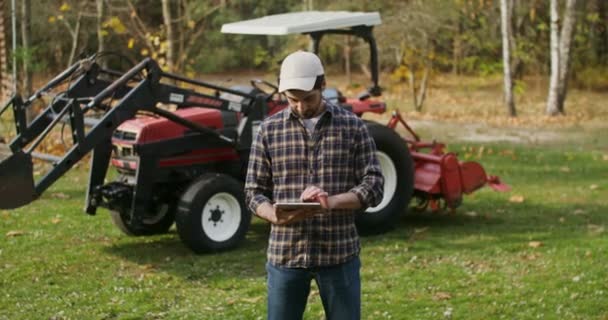 Een jonge man gebruikt een tablet, die op herfstdag bij een landbouwmachine staat — Stockvideo