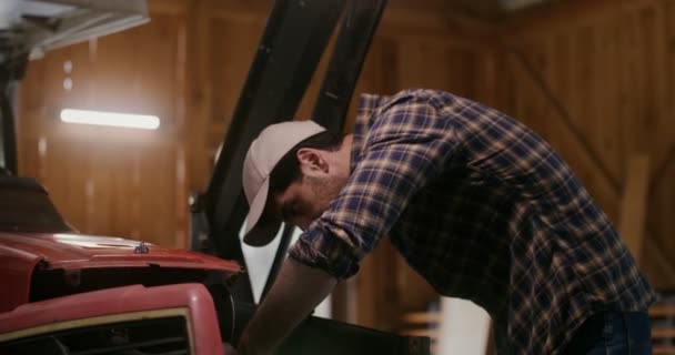 Een jongeman repareert een kleine landbouwtrekker in een houten schuur — Stockvideo