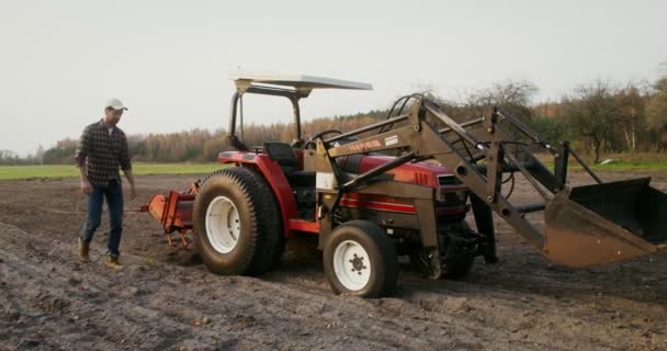 Un hombre camina a través de un campo arado a un tractor y se sienta en el asiento en la cabina — Vídeo de stock