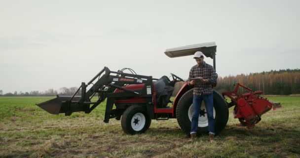 年轻的拖拉机司机靠在一辆现代农用拖拉机上使用平板电脑 — 图库视频影像
