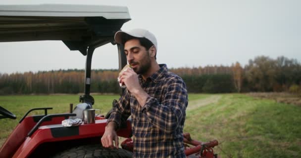 Joven agricultor comiendo un sándwich mientras está de pie cerca de una moderna máquina agrícola — Vídeos de Stock